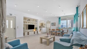 Un salon moderne avec un canapé, une table basse et un téléviseur. Il y a également un coin repas avec des chaises et une table. Les murs de la pièce sont blancs et le sol est carrelé.