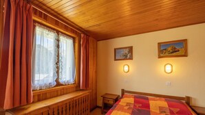 A wooden-paneled room with a bed, a window with curtains, a nightstand, and two wall-mounted lights. - Apartment 'La Rose des Vents N 1' with Mountain View, Balcony & Wi-Fi (Châtel)