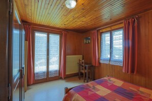 Een kamer met houten wanden en plafond, een bed met een beddeken met dessin, een bureau en een stoel.