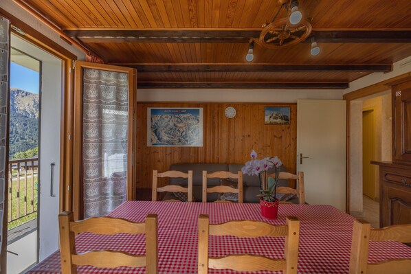 A dining area with a checkered tablecloth, wooden chairs, a wooden ceiling, and a view of mountains through a door.