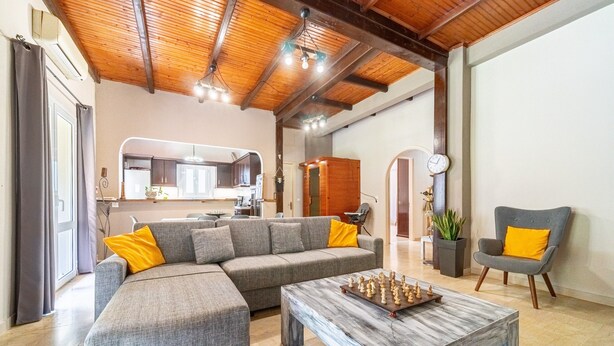 Een woonkamer met een modulaire bank, een salontafel met schaakstukken, een grijze fauteuil, en een keuken op de achtergrond.