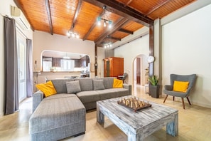 A living room with a sectional sofa, a coffee table with chess pieces, a grey armchair, and a kitchen in the background.
