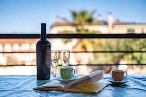 Une table avec un livre, deux tasses et une bouteille, placée sur un balcon doté d’une balustrade et offrant une vue sur des immeubles et des arbres.