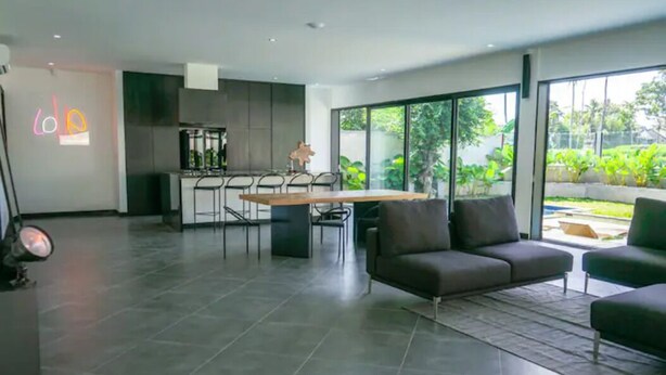 Una sala de estar moderna con un ventanal grande, un sofá, una zona de comedor con sillas y una cocina al fondo.