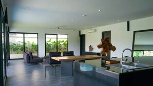 Una cocina moderna con una isla grande, un comedor con sofá y sillas, y ventanas amplias con vistas a la vegetación.