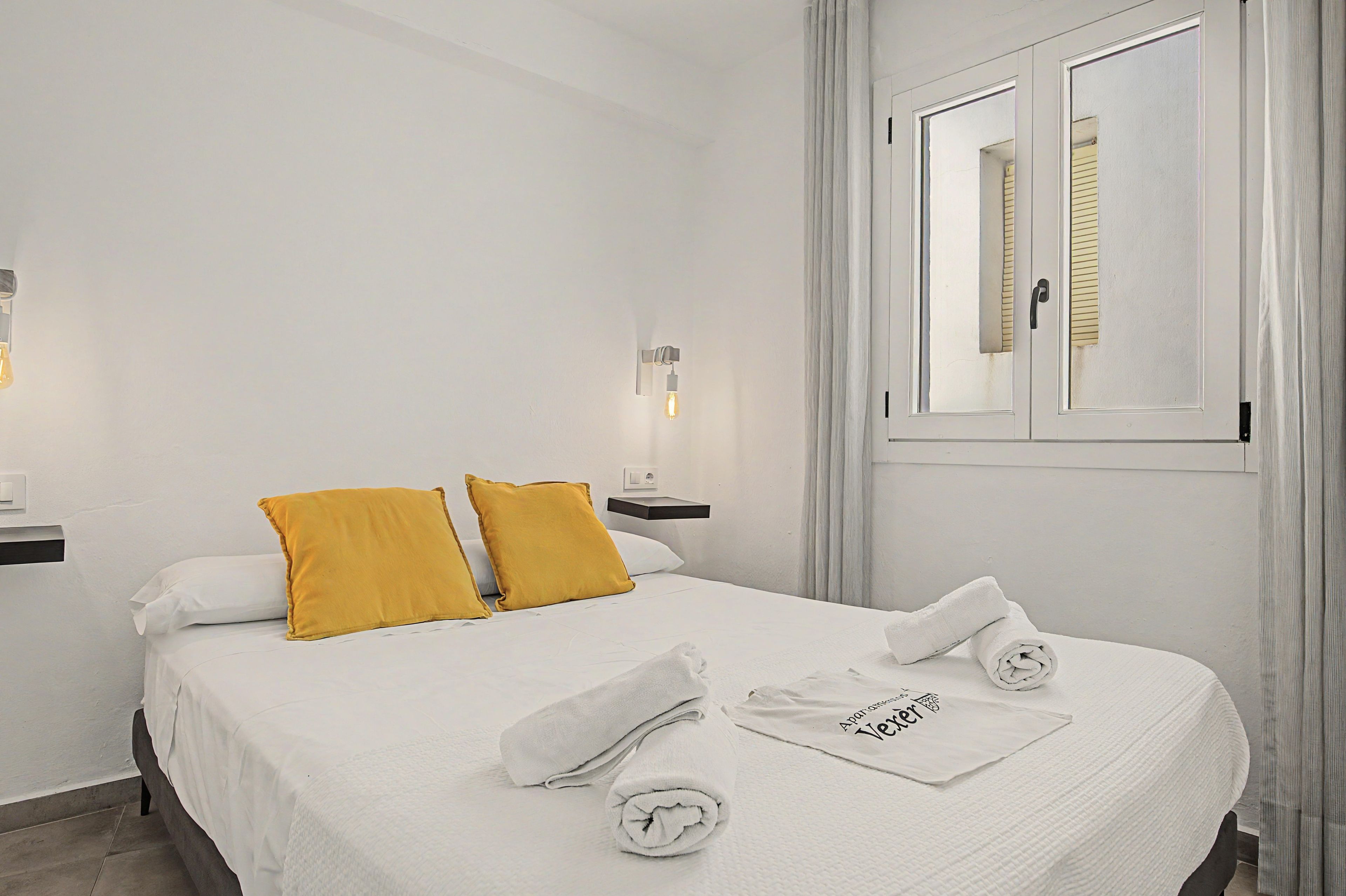 A hotel room with a bed, two pillows, a nightstand, a lamp, a window with curtains, and a towel with text.