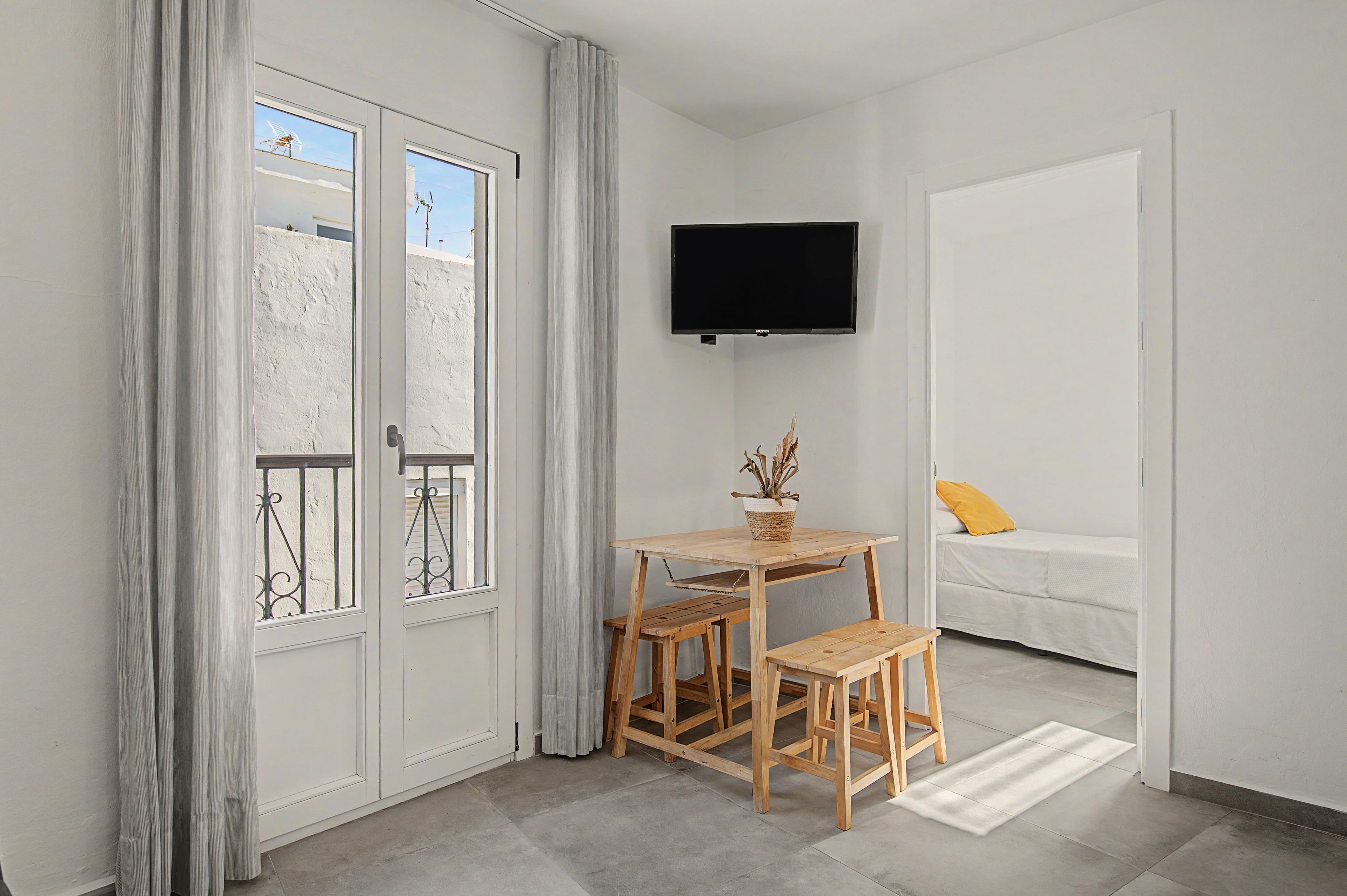 A small room with a wooden table and chairs, a bed with a yellow pillow, a mounted TV, and a window with curtains.