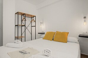 A hotel room with a bed, a wooden wardrobe, a wall-mounted light fixture, and a bedside table with a power outlet.