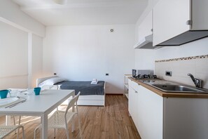A compact room with a bed, a small kitchenette, a dining area, and a white wall with a light switch.