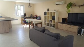 Ein modernes Wohnzimmer mit grauem Sofa, einer Essecke mit weißem Tisch und Stühlen, einem Fernseher auf einer Holzkommode und einer eingebauten Bücherreihung.