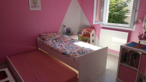 Ein Kinderzimmer mit einem Einzelbett, einem kleinen Schreibtisch, einem Bücherregal und einem Fenster mit Blick auf Grünflächen.