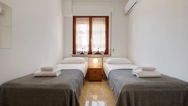 Ein Hotelzimmer mit zwei Einzelbetten, einem Nachttisch mit Lampe, einem Fenster mit Vorhängen und einer Klimaanlage.