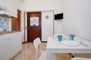 A kitchen with a white countertop, a sink, and a stove. There is a dining table with a white plate, blue cups, and cutlery. A clock is on the wall, and a door with a sign is visible.