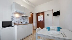 Eine kleine Küche mit Mikrowelle, Spüle und einem Fliesenspiegel mit Muster. Außerdem sind ein Esstisch mit Stühlen und ein an der Wand befestigter Fernseher zu sehen.