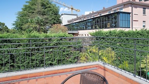 Ein Balkon mit Metallgeländer, einem Stuhl und Blick auf ein Gebäude mit Glasfassade sowie einem Baukran im Hintergrund.