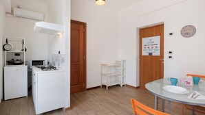 A compact kitchen with a stove, microwave, and white cabinets. There is a dining area with a glass table and orange chairs. - Apartment Civico22 - Bilo 3 with Balcony, A/C & Wi-Fi (Peschiera del Garda)