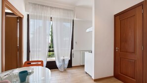A room with a dining table, chairs, a window with sheer curtains, a wooden door, and a radiator. - Apartment Civico22 - Bilo 3 with Balcony, A/C & Wi-Fi (Peschiera del Garda)