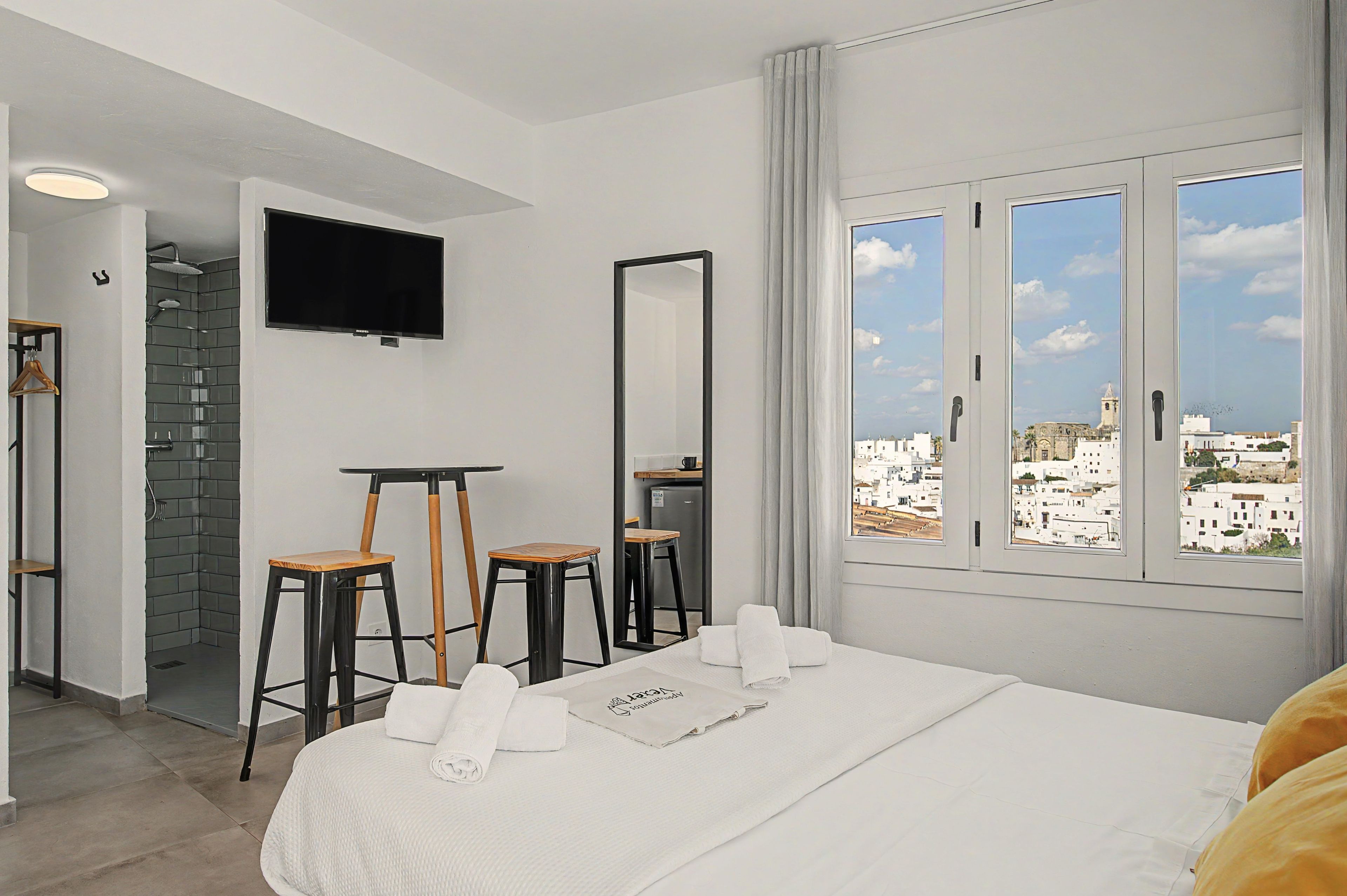 Een hotelkamer met een bed, twee krukken, een televisie, een spiegel en een raam met uitzicht op de stad.