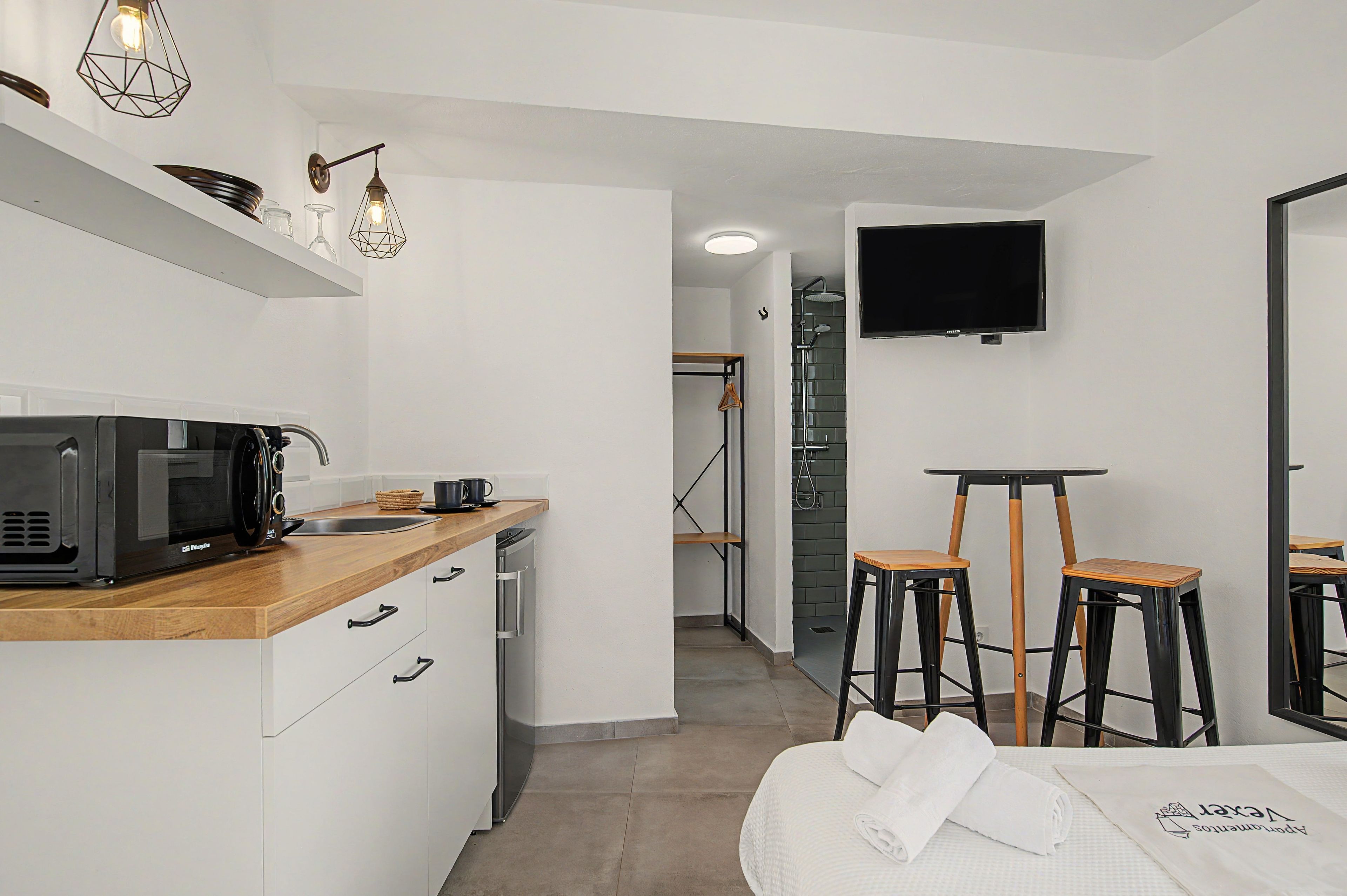 A modern kitchen with a microwave, coffee maker, and wooden countertop. There is a small seating area with two stools and a table, and a TV mounted on the wall.