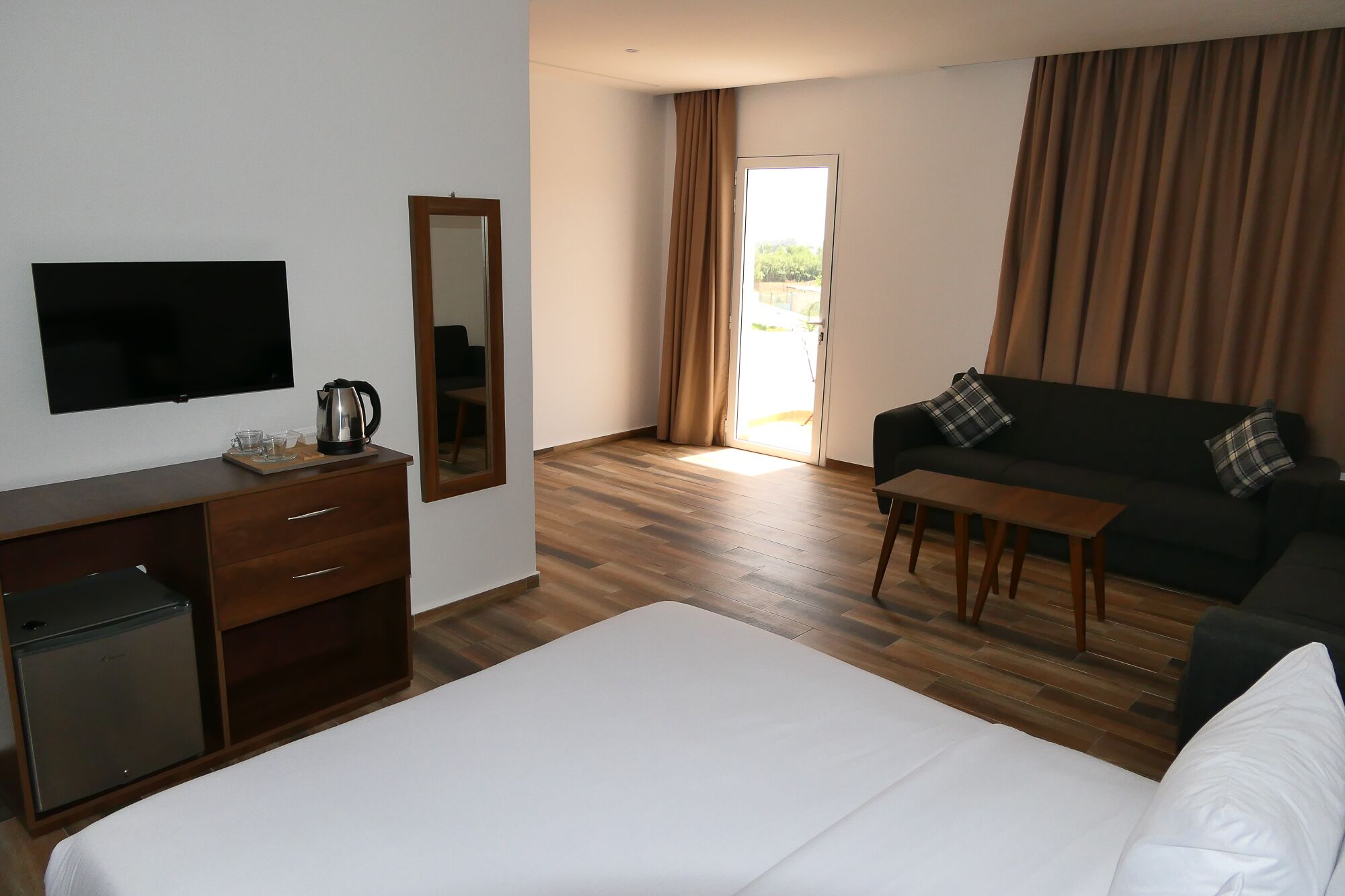 Une chambre d’hôtel équipée d’un lit, d’une télévision, d’un bureau avec une bouilloire, d’un canapé et d’une porte-fenêtre coulissante donnant sur l’extérieur.