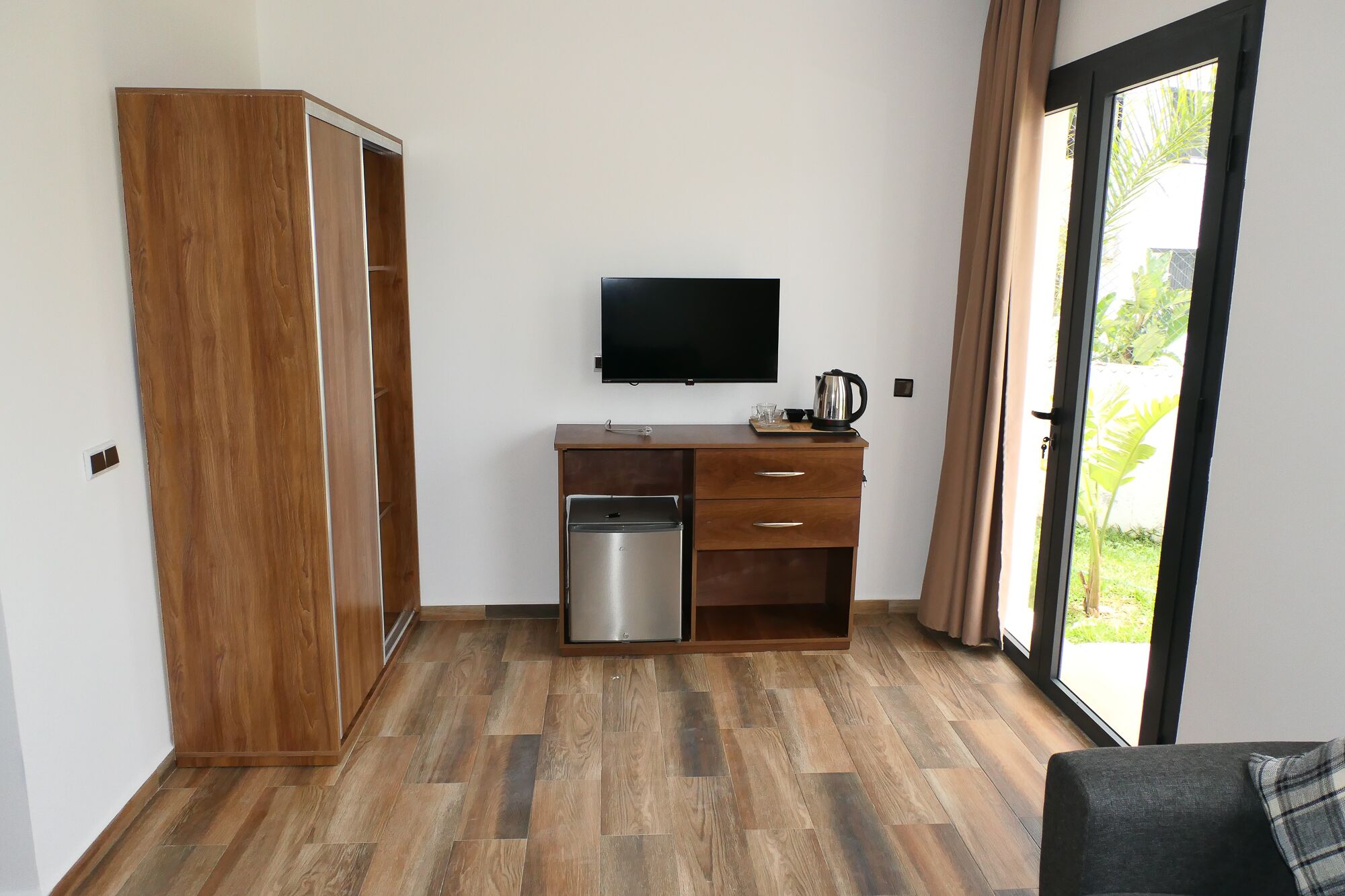 A room with a wooden wardrobe, a kitchenette with a sink and a stove, a flat-screen TV mounted on the wall, and a sliding glass door leading to an outdoor area.