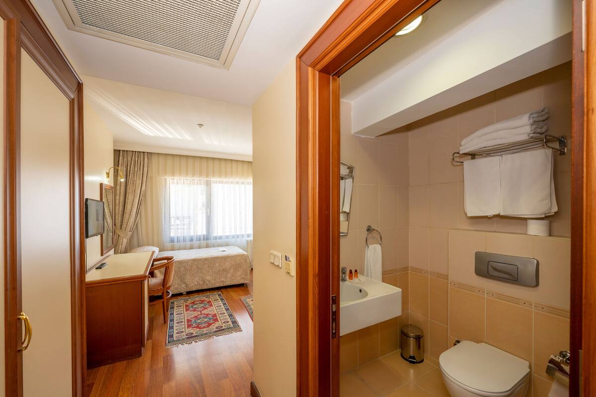 Une chambre d’hôtel avec un bureau en bois, un lit recouvert d’une courtepointe beige, une salle de bain équipée d’un lavabo, de toilettes et d’un porte-serviettes.