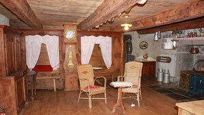 Ein rustikaler Holzraum mit Holzboden, zwei Korbstühle mit roten Kissen, ein Holztisch, eine Uhr an der Wand und ein Steinkamin mit Metalltöpfen.