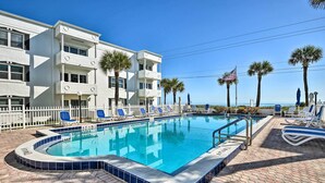A swimming pool with lounge chairs, a white building with balconies, and palm trees. - Coastal Condo Escape: Steps to Beach (Ormond Beach)