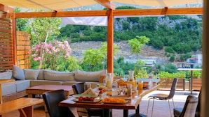 Un espacio exterior cubierto para comer con mesa de madera preparada para el desayuno, un sofá y vistas a un paisaje montañoso.