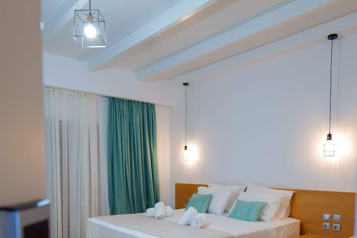 A modern hotel room with a wooden floor, a large bed with white linens, teal and white pillows, and two pendant lights.