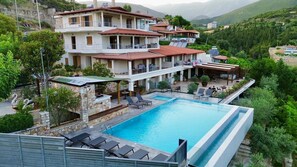 A multi-story hotel with a swimming pool, outdoor seating, and a mountainous backdrop. - Royal 1967 (Himarë)