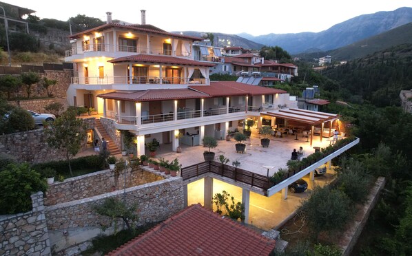 Una villa de varios pisos con terraza, muros de piedra y un paisaje montañoso de fondo.