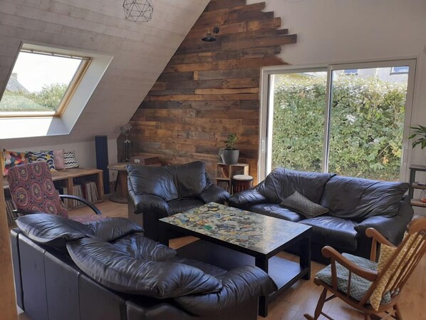 A modern loft with a sloped ceiling, a black leather sofa set, a coffee table with newspapers, and a large window with a view of greenery.