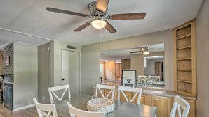 Un comedor con mesa de madera y sillas blancas, ventilador de techo y cocina al fondo.