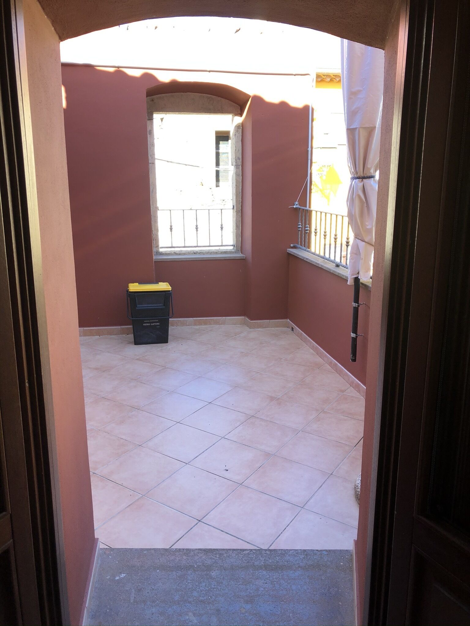 A small, enclosed balcony with a tiled floor, a window, and a black trash bin.