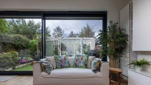 A modern living room with a white sofa, colorful tropical-themed cushions, a wooden floor, and a large glass door leading to a garden. - Luxe 3 Bed property with beautiful garden room. (Harrogate)