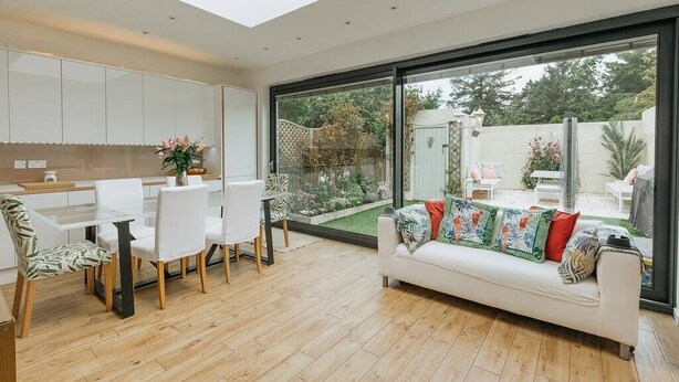 A modern kitchen with white cabinets, a dining area with white chairs, and a large sliding glass door leading to a garden. - Luxe 3 Bed property with beautiful garden room. (Harrogate)