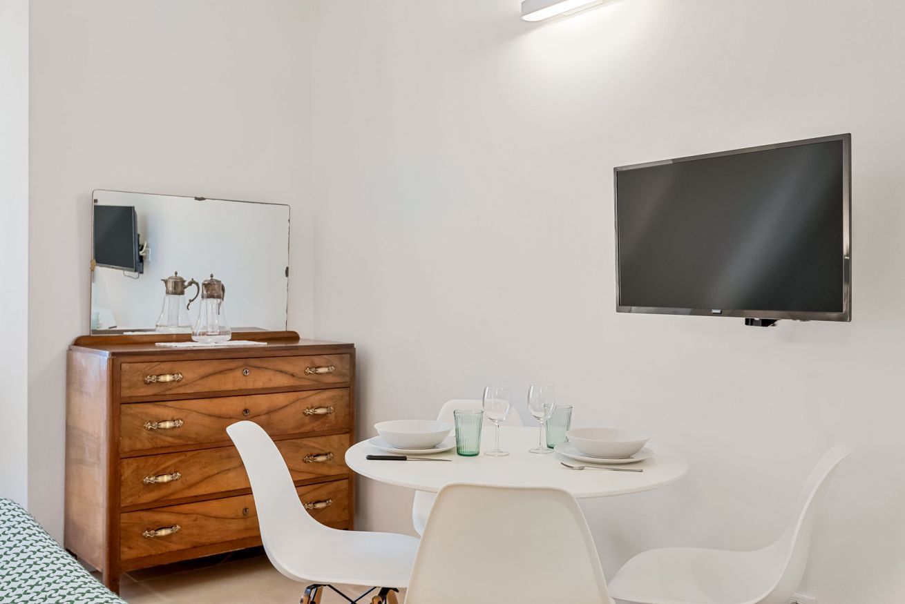 A modern room with a wooden dresser, a round dining table with chairs, a flat-screen TV mounted on the wall, and a mirror above the dresser.