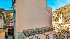 A building with a balcony, a staircase, and a view of a town with buildings and a church. - GuestHost - Riomaggiore cozy studio x4! (Riomaggiore)