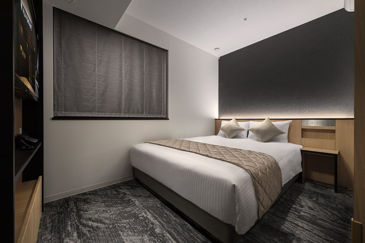 Une chambre d’hôtel moderne dotée d’un grand lit, d’une table de chevet en bois, d’une étagère intégrée et d’une grande fen être avec des stores.