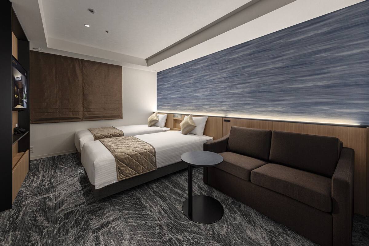 Une chambre d’hôtel moderne, dotée d’un grand lit, d’un canapé, d’une petite table ronde et d’une grande fenêtre offrant une vue sur le ciel.