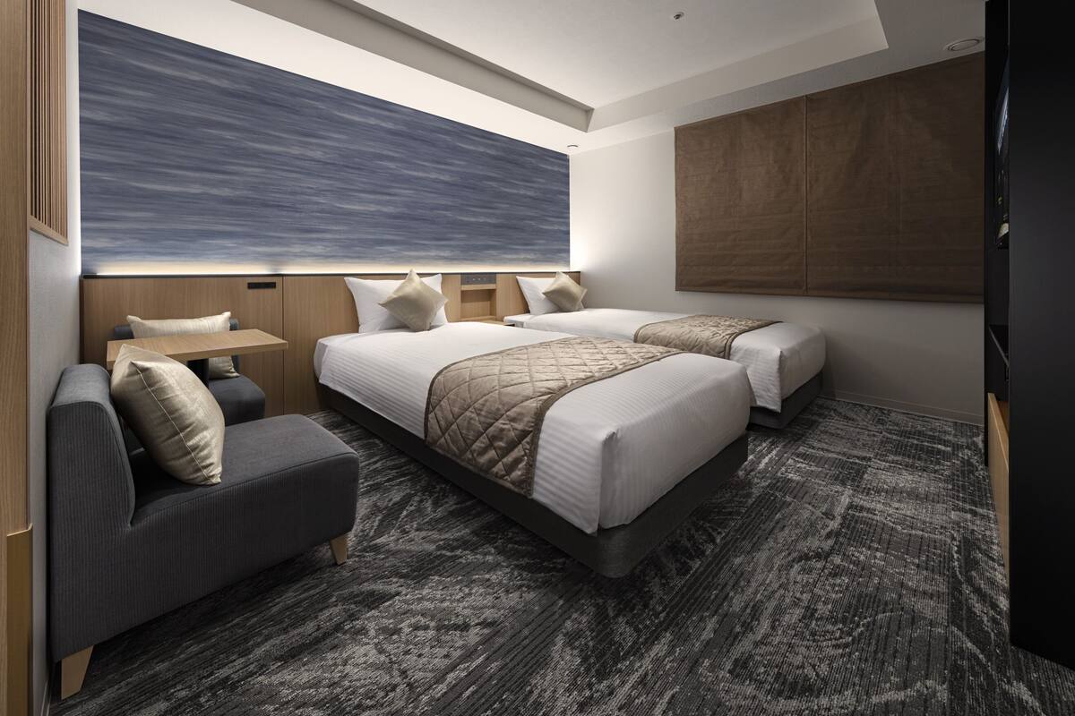 Une chambre d’hôtel moderne avec deux lits, un canapé, un grand tableau abstrait au mur et une moquette à motifs.