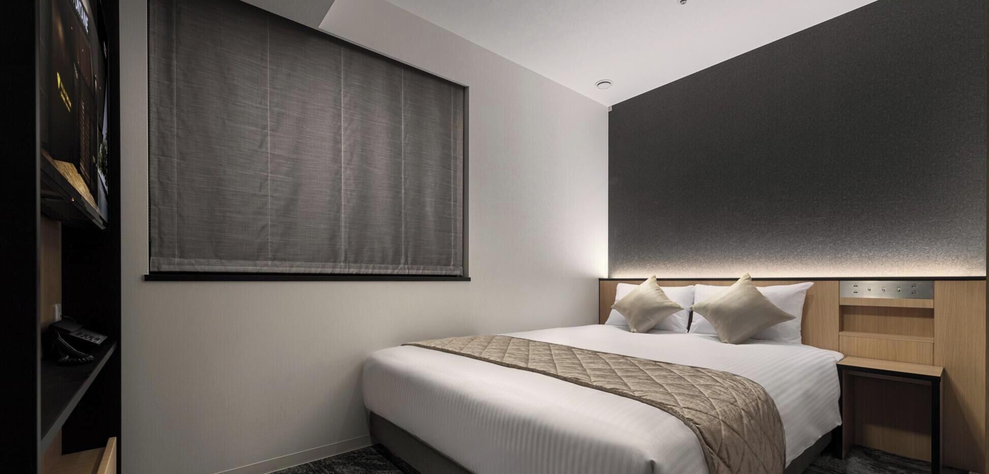 A hotel room with a bed, a large window with a blind, a headboard with built-in lighting, and a small bedside table.