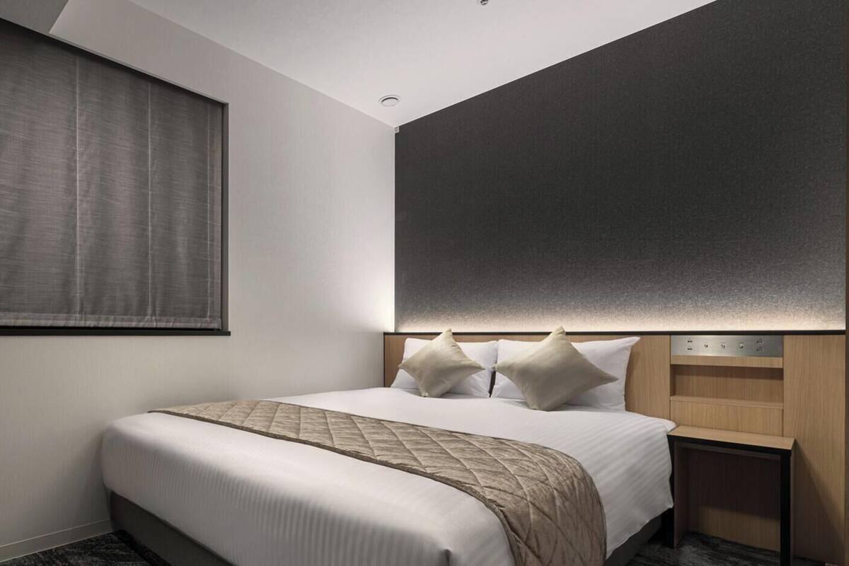 Une chambre d’hôtel comprenant un lit, une grande fenêtre avec un store, une tête de lit dotée d’un éclairage intégré et une petite table de chevet.