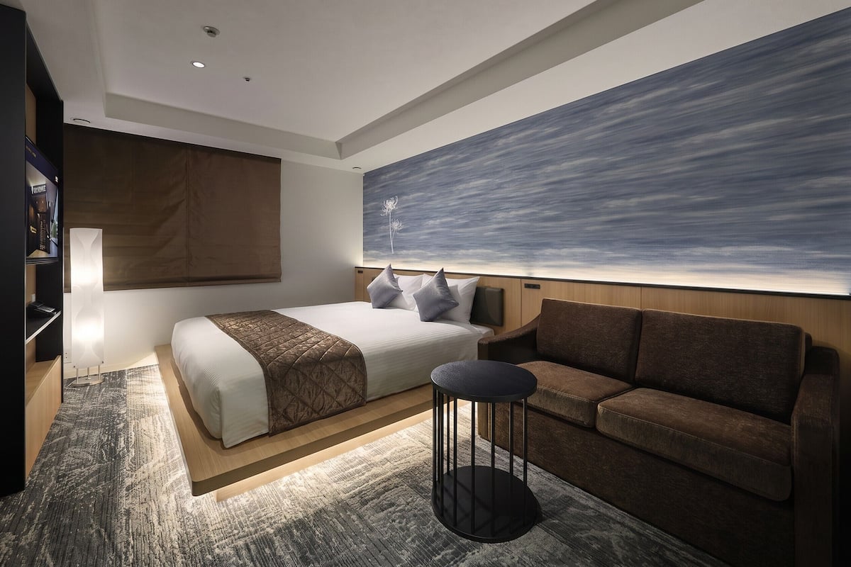 Une chambre d’hôtel moderne avec un grand lit, un canapé, une petite table ronde et un grand tableau abstrait accroché au mur.
