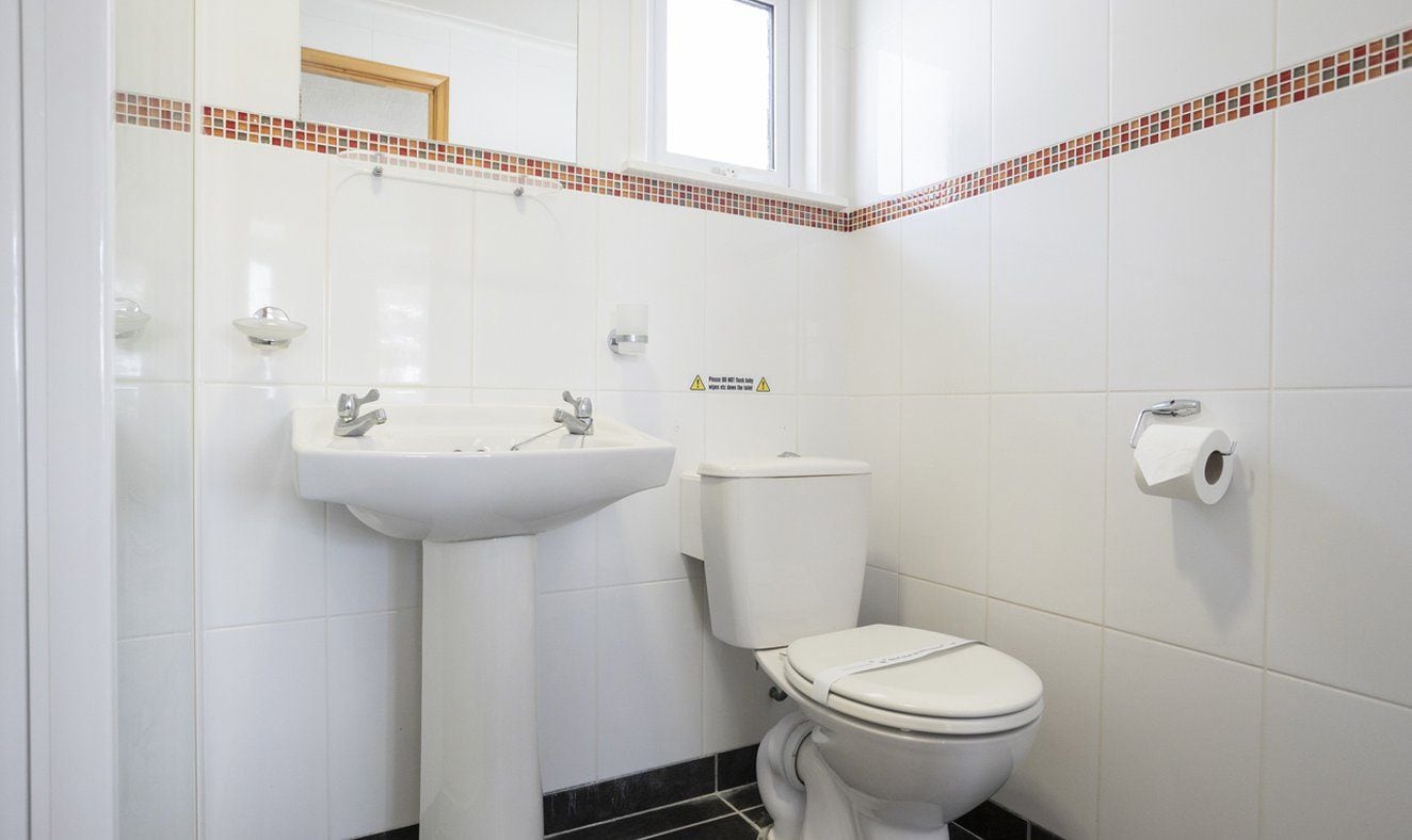 Une salle de bain avec un lavabo blanc, des toilettes et un miroir. Il y a une fenêtre au-dessus du lavabo et une bordure de carreaux qui entoure le sol.