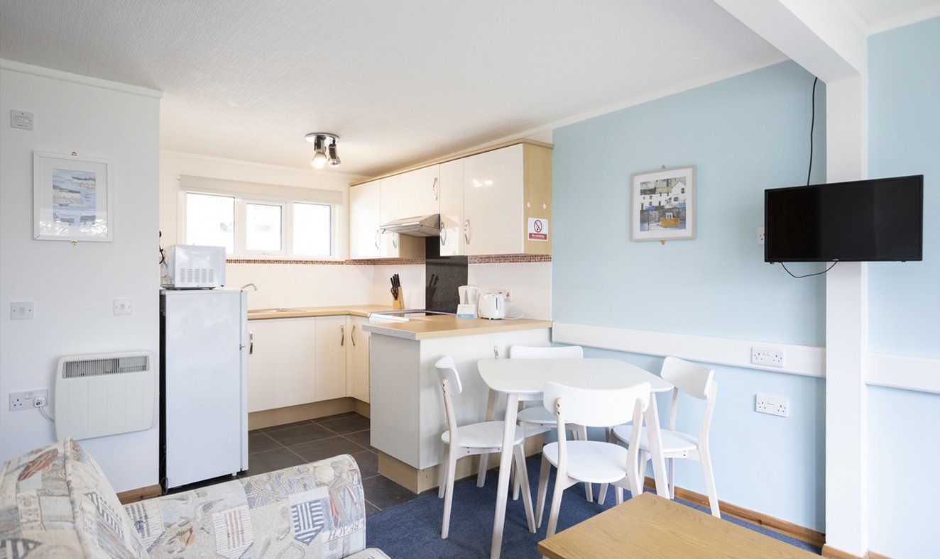 Een kleine keuken met witte kastjes, een magnetron en een koelkast. Er is een zithoek met een tafel en stoelen.