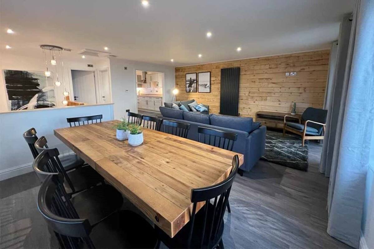 A modern living space with a wooden dining table, black chairs, a blue sofa, and a kitchen in the background.