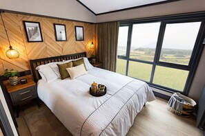 A bedroom with a large bed, a nightstand, a lamp, and a view of the outdoors through a window.