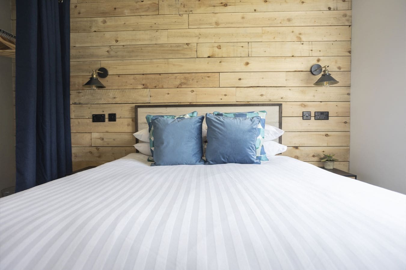 Een bed met een gestreept wit dekbed, twee blauwe kussens en een houten hoofdbord.